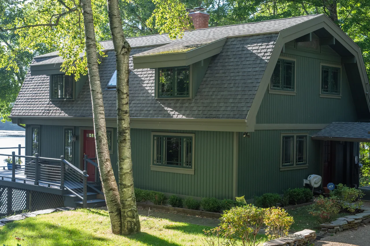 Sage green house with a dark shingled roof, elevated deck and surrounding trees